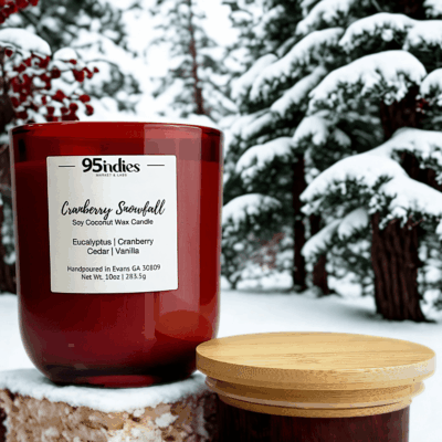 Cranberry Snowfall candle in a red glass jar with snowy pine trees and berries in the background.