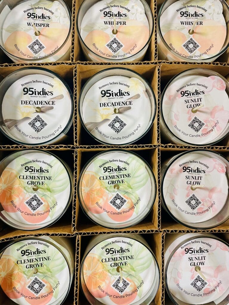 Box of twelve 95indies candles in assorted fragrances, including Whisper, Decadence, Clementine Grove, and Sunlit Glow, displayed in individual glass tumblers with branded dustcovers