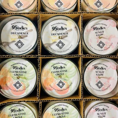 Box of twelve 95indies candles in assorted fragrances, including Whisper, Decadence, Clementine Grove, and Sunlit Glow, displayed in individual glass tumblers with branded dustcovers