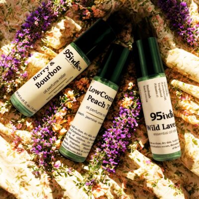 A top-down photo of three 95ndies perfume rollers on a floral cloth background with fresh lavender sprigs. The roller on the left is "Berry & Bourbon," the middle is "LowCountry Peach Tea," and the right is "Wild Lavender." Each bottle is clear glass with a black cap.