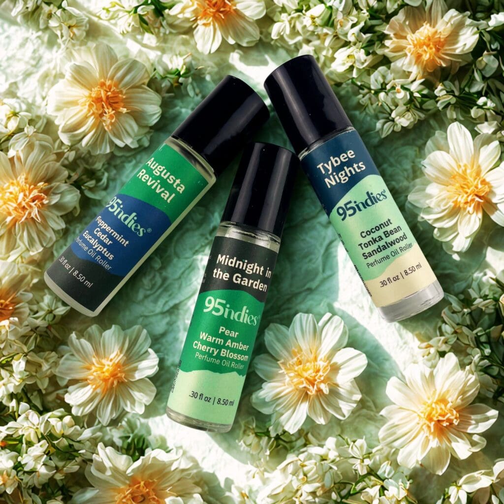 Alt Text A top-down photo of three 95ndies perfume rollers arranged on a light green fabric surrounded by white and yellow flowers. The rollers are labeled "Augusta Revival," "Midnight in the Garden," and "Tybee Nights."