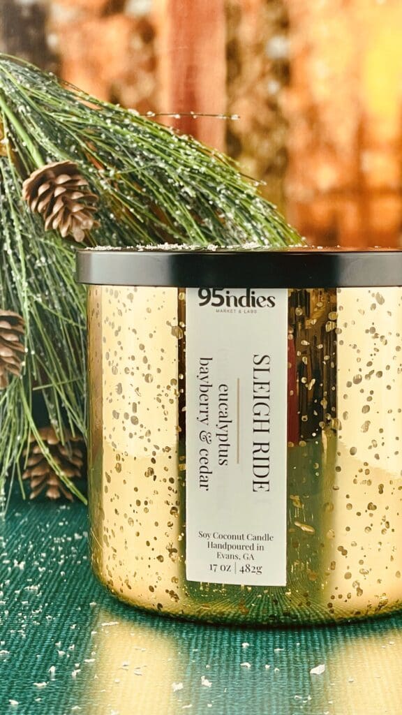 Gold mercury glass candle labeled “Sleigh Ride” by 95indies, surrounded by evergreen branches and pinecones.