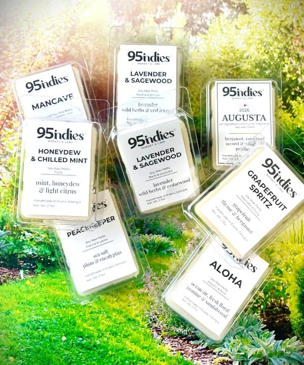 Assorted 95indies soy wax melt clamshells displayed on a sunlit garden background, featuring fragrances including Lavender & Sagewood, Honeydew & Mint, Grapefruit Spritz, Augusta, Aloha, and Peacemaker.